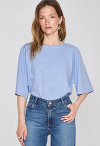Maglietta azzurra chiaro a maniche corte con scollatura tonda e dettaglio di cucitura triangolare. Indossata con jeans in denim a vita alta. Tessuto morbido.
