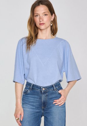 Light blue short-sleeve top with a rounded neckline and triangular seam detail. Worn with high-waisted denim jeans. Soft fabric texture.