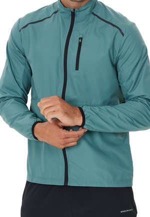 Hombre vistiendo chaqueta con cremallera color verde azulado con cremallera negra y bolsillo en el pecho, ajustándose la manga, combinado con pantalones negros etiquetados como "Endurance".