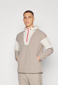 Lightweight, gray and cream jacket with a hood, half-zip front, side pockets, and red accents. Textured fabric with contrasting panels.