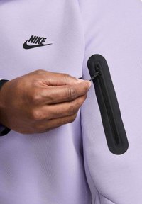 Felpa Nike di colore viola chiaro con logo nero e tasca con zip sulla manica sinistra, caratterizzata da una texture di tessuto morbido.