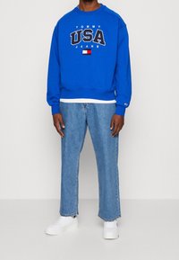 Modrá mikina s nápisem "TOMMY USA JEANS", s bílým kulatým výstřihem a pružným lemem. Kombinována s světle modrými džíny a bílými teniskami.