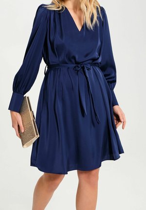 Kvinde iført en mørkeblå langærmet V-hals kjole med bindebånd i taljen, som holder en beige tekstureret clutch i den ene hånd.