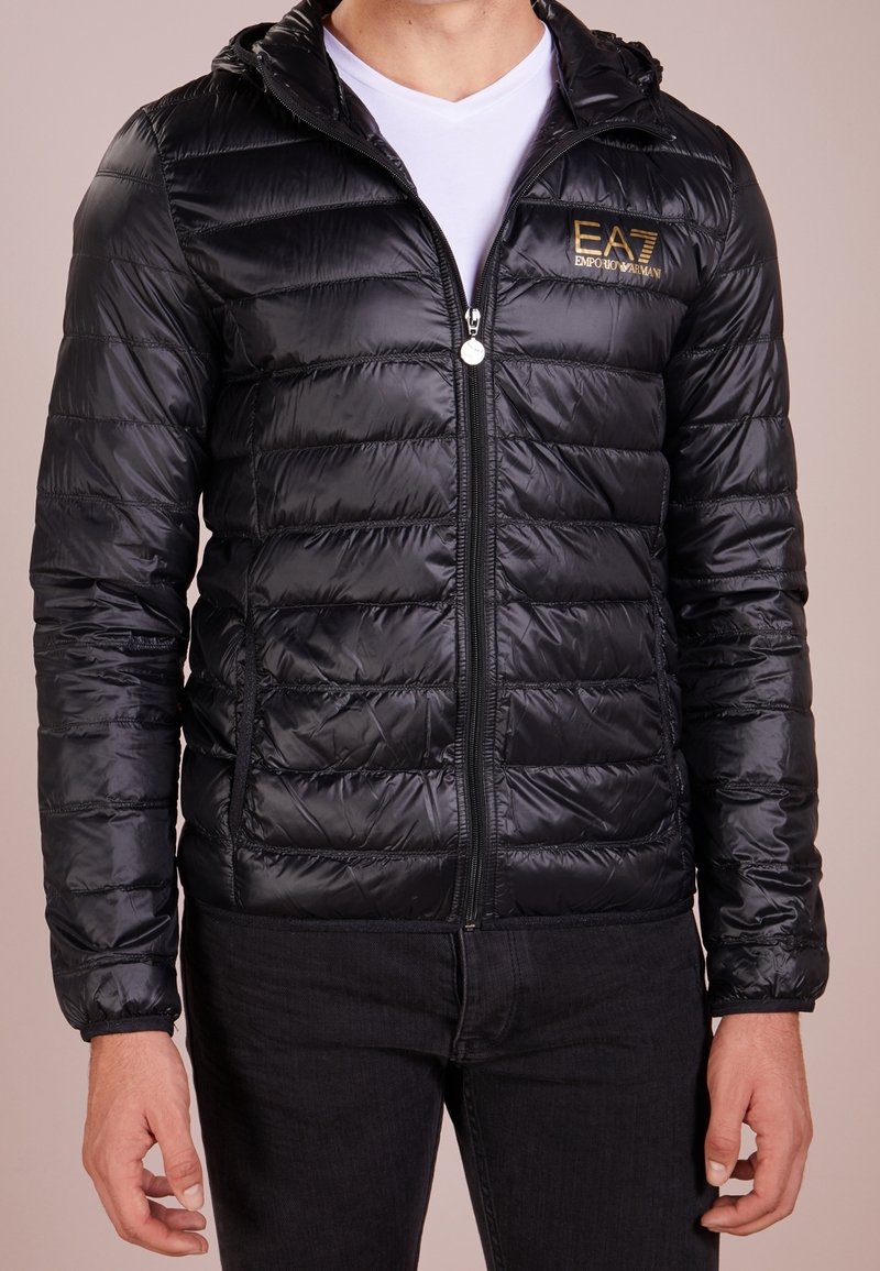 Homme portant une veste matelassée noire zippée avec capuche, t-shirt blanc en dessous et pantalon noir, debout devant un fond neutre.