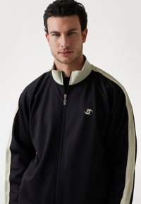 Veste zippée noire avec des accents beige clair le long des épaules, présentant un petit logo sur la poitrine gauche et un col montant.
