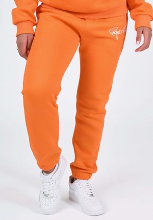 Persona che indossa pantaloni della tuta arancioni vivaci con il logo "Project X" sulla coscia e sneakers bianche, in piedi davanti a uno sfondo bianco semplice.