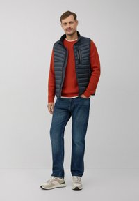 Marineblauer Steppweste über einem orangefarbenen Pullover, blaue Jeans und beigen Sneakers mit grünen Akzenten. Die Weste hat eine Reißverschlusstasche.