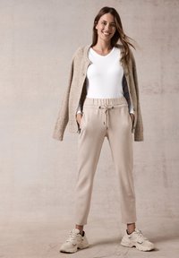 Beige jogger-housut, joissa on elastinen vyötärö, etutaskut ja nyöritys. Yhdistetty valkoisen V-paitan ja beige neulotun neuletakin kanssa.