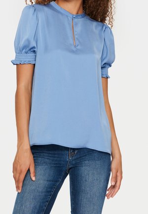 Kobieta ubrana w jasnoniebieską bluzkę z krótkim rękawem, z marszczonymi mankietami i małym wycięciem przy szyi zapinanym na guziki, w połączeniu z niebieskimi dżinsami.