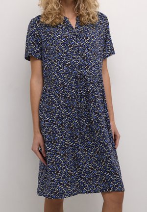 Kvinde iført en knælængde kjole med korte ærmer og et abstrakt blomstermønster i blå, hvid og sort mod en ensartet lys baggrund.