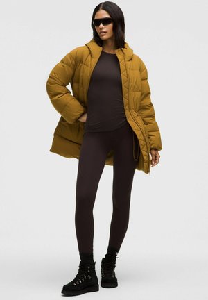 Puffjakke i sennepsgul, tætsiddende brun top, sorte leggings og sorte støvler med snørebåndsdesign. Mat overflade og afslappet pasform.