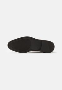 Suela de goma negra con agarre texturizado, forma curva y ligero tacón. Parte superior de cuero marrón claro con un acabado suave y costuras mínimas.