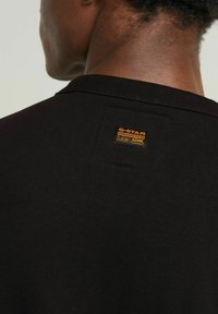 T-shirt nera in cotone con texture a costine, dotata di un'etichetta quadrata sul retro con testo e logo dorati. Design semplice e minimalista.