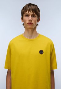 Ung person med kort brunt hår iført en enkel gul t-shirt med et lille rundt sort og orange logo på venstre bryst.