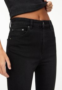 Vaqueros de mezclilla negra con cintura alta, diseño de cinco bolsillos y acentos de metal. Textura suave con una silueta ajustada.