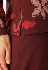 Bordeaux top met bloemenprint in rood en roze, met een splitrand en zachte katoen textuur. Close-up van een hand die de stof vasthoudt.
