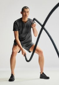 Sort tekstureret atletisk T-shirt og grå shorts. Personen griber en tyk sort battle rope med sorte atletiske sko på en lys baggrund.