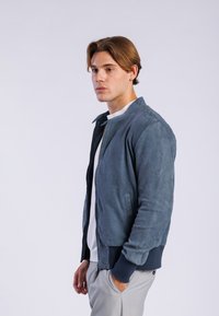 Chaqueta bomber de ante en azul profundo, con cierre de cremallera, puños acanalados y bolsillos laterales, combinada con una camisa clara y pantalones grises.