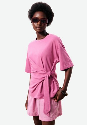 Femme avec afro court portant un haut rose surdimensionné noué à la taille, un short rose à boutons, de grandes lunettes de soleil foncées et un bracelet épais.
