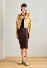 Veste en suède beige courte avec zips, robe midi brune à côtes, et bottines beiges à semelles épaisses. Écharpe avec des motifs abstraits.