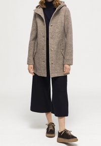 Cappotto di pile beige con cappuccio, bottoni frontali e tasche laterali, abbinato a pantaloni neri cropped e scarpe marroni con lacci e suola strutturata.