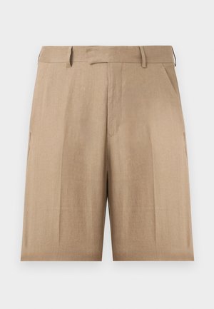 Bărbat purtând o cămașă bleu cu mâneci lungi cu nasturi, pantaloni scurți bej până la genunchi, șosete albe și adidași albi, stând cu mâinile în buzunare.