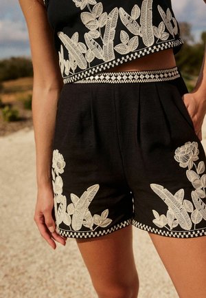 Femme portant un short noir avec une broderie florale blanche et un crop top assorti, se tenant à l'extérieur sur un chemin de gravier.