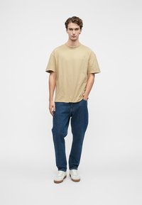 Beige bavlněné tričko s kulatým výstřihem a krátkými rukávy, spárované s tmavě modrými džíny. Bílé tenisky s hnědými podrážkami. Jednoduchý design.