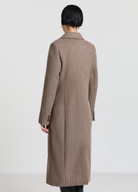Lungo cappotto di lana in tessuto marrone strutturato, con collo a rever e polsini con bottoni. Sul retro si distingue una silhouette sagomata e tasche laterali.