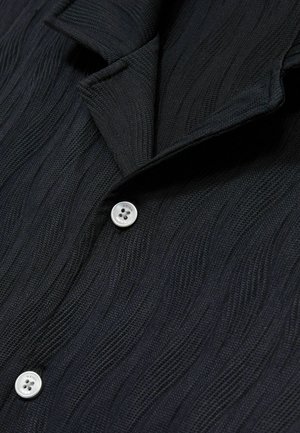 Zwarte shirt met textuur en golfpatroon, voorzien van een zilveren knop met het etiket "REISS" bij de kraag. De stof is gebreid en zacht.