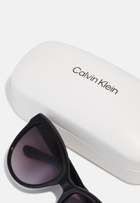 Črne sončne očala z gradientnimi lečami, postavljena ob beli trdi etui, označen z napisom "Calvin Klein" v črni pisavi.