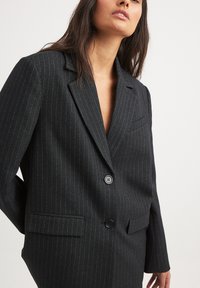 Blazer noir à rayures avec un col à cran, fermeture à un bouton et poches avant. Fabriqué en tissu texturé avec un éclat subtil.