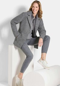 Lächelnde Frau in gestapeltem grauen Outfit und weißen Sneakers posiert sitzend auf weißen geometrischen Blöcken vor einem einfarbigen Hintergrund.