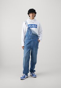 Jeansin overall med én stropp, lyseblå farge, løs passform. Hvit genser med blå "HUGO"-logo. Sportssko med blå og hvite detaljer.
