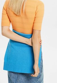 Pletený top v oranžové barvě s žebrovanou strukturou, kombinovaný s modrým zavinovacím pláštěm, se žebrovaným lemem. Nosit na džínách, ukazuje vrstvené barvy.