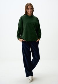 Felpa verde in velluto con vestibilità rilassata e tasca frontale, abbinata a pantaloni in velluto blu navy con tasche laterali e una vita comoda.