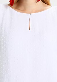 Blusa bianca in tessuto testurizzato con punti in rilievo, apertura a chiave sul collo e piccolo bottone metallico decorativo.
