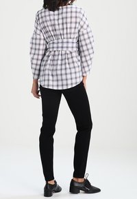 Camicia a quadri bianca e viola con vita aderente, maniche lunghe e un dettaglio di laccio sul retro, abbinata a pantaloni skinny neri e scarpe nere.