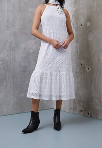 Femme portant une robe mi-longue blanche en dentelle sans manches, des bottines noires et une écharpe noire et blanche, debout contre un mur en béton.
