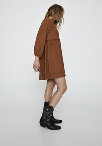 Rostfarbendes, langärmeliges Kleid mit gestuftem Design und strukturiertem Stoff. Kombiniert mit schwarzen Leder-Cowboy-Stiefeln mit dekorativer Naht.