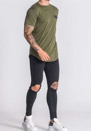 Homme portant un t-shirt vert olive, un jean skinny noir déchiré, des baskets blanches, se tenant debout avec des tatouages visibles sur les bras sur un fond uni.