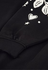 Zwarte sweatshirtmouw met witte geborduurde ontwerpen, waaronder bladeren, harten, sterren en cirkelvormige accenten. Zacht, gestructureerd materiaal met geribbelde manchetten.