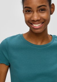 Teal-gestreiftes T-Shirt mit Rundhalsausschnitt und kurzen Ärmeln, die einen gewellten Rand haben, getragen von einer Person vor einem neutralen Hintergrund.