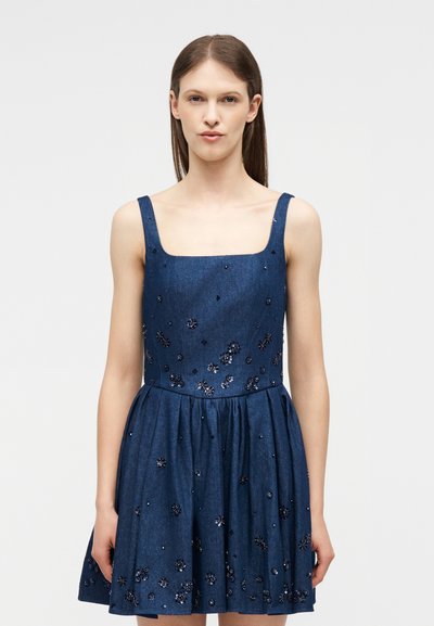 Γυναίκα που φοράει αμάνικο μπλε φόρεμα από τζιν με διακοσμητικά λουλούδια από χάντρες, στέκεται μπροστά σε απλό φωτεινό φόντο.