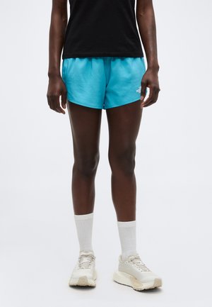 Személy élénk kék sport rövidnadrágban, fekete pólóban, fehér bokazokniban és törtfehér sportcipőben, tiszta háttér előtt áll.