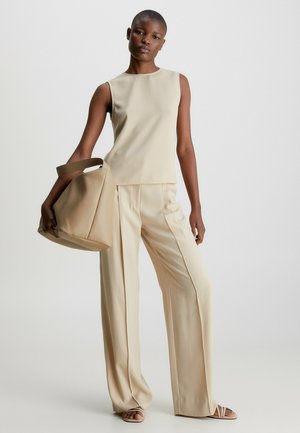 Top sans manches beige et pantalon large, fabriqués en tissu lisse. Le modèle tient un sac à main assorti en doux avec une bandoulière.