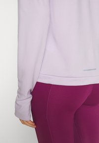 Maglietta lunga di colore viola chiaro abbinata a leggings scuri viola a vita alta. La maglietta presenta un accento riflettente vicino all'orlo.