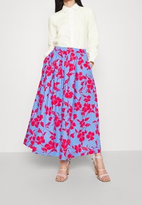 Gonna maxi floreale in cotone azzurro chiaro, con grandi fiori rossi, una vita elastica e tasche laterali. Abbinata a una camicia bianca con colletto.