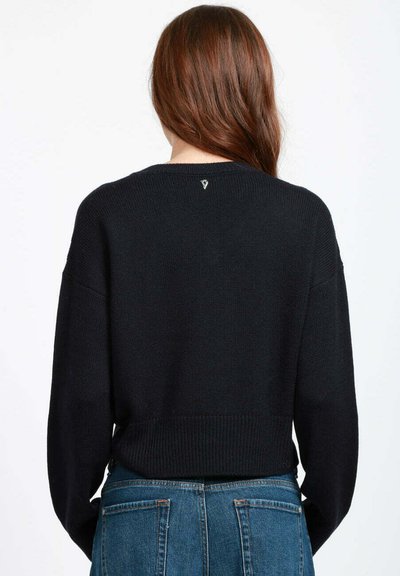 Donna con i capelli lunghi e castani che indossa un pullover nero di maglia e jeans blu, vista di spalle su uno sfondo bianco.