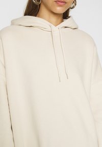 Sudadera beige con textura suave, cordones en la capucha y hombros caídos. Sin patrones ni detalles de hardware visibles.